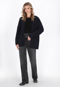 Cardigan fuzzy blu navy con apertura frontale, maniche lunghe e tasche, abbinato a una maglietta nera aderente e jeans grigi a vita alta.