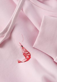 Sweat à capuche rose avec cordons présentant une crevette rouge brodée en détail près du poignet côtelé.