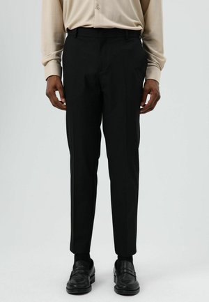 Zwarte, op maat gemaakte broek met een smalle pasvorm, voorzien van twee zijzakken en een gladde textuur. Draagt een beige overhemd met knopen en loafers.