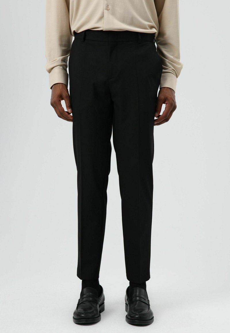 Zwarte, op maat gemaakte broek met een smalle pasvorm, voorzien van twee zijzakken en een gladde textuur. Draagt een beige overhemd met knopen en loafers.