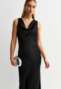 Abito nero senza maniche con scollo a cowl, texture liscia, orlo asimmetrico e leggero drappeggio. Una pochette circolare argentata in mano.