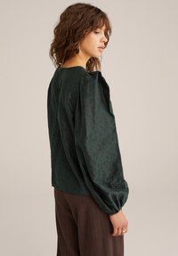 Blouse longue à manches longues vert foncé en tissu doux et texturé, avec des manches bouffantes et un col rond. À associer avec un pantalon à rayures brunes.
