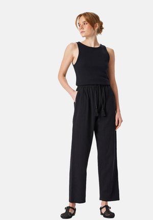 Kvinde iført en sort ærmeløs top, højtaljede brede bukser med snor i taljen og sorte vævede sandaler, stående med den ene hånd i lommen.