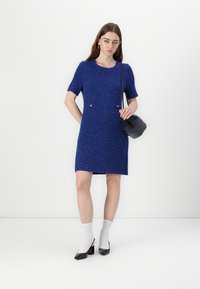 Korte mouwen, jurk tot de knie in gestructureerde blauwe stof met decoratieve knopen bij de taille. In combinatie met een kleine zwarte tas en schoenen.