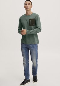 Langarm grünes T-Shirt mit einer dunkelgrünen Brusttasche und Reißverschlussdetails, kombiniert mit hellblauen Jeans und schwarzen Sneakern.