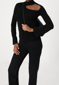 Schwarzes langärmliges Oberteil mit einem Stehkragen und einem Ausschnitt an einer Schulter, kombiniert mit schwarzen Hosen und einer Tasche. Glatte Stofftextur.