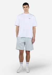 Camiseta blanca de algodón con un pequeño logo negro en el pecho, combinada con pantalones cortos a rayas ligeras en color azul y zapatillas blancas.