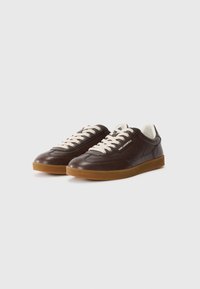 Sneaker in pelle marrone con suole in gomma beige chiaro, dotate di lacci crema e marchio sul lato. Texture liscia con un design classico.