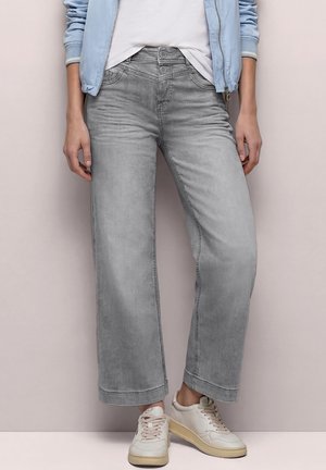 Persoon met lichtblauwe jas, wit shirt, grijze brede jeans en witte sneakers, staand tegen een effen achtergrond.