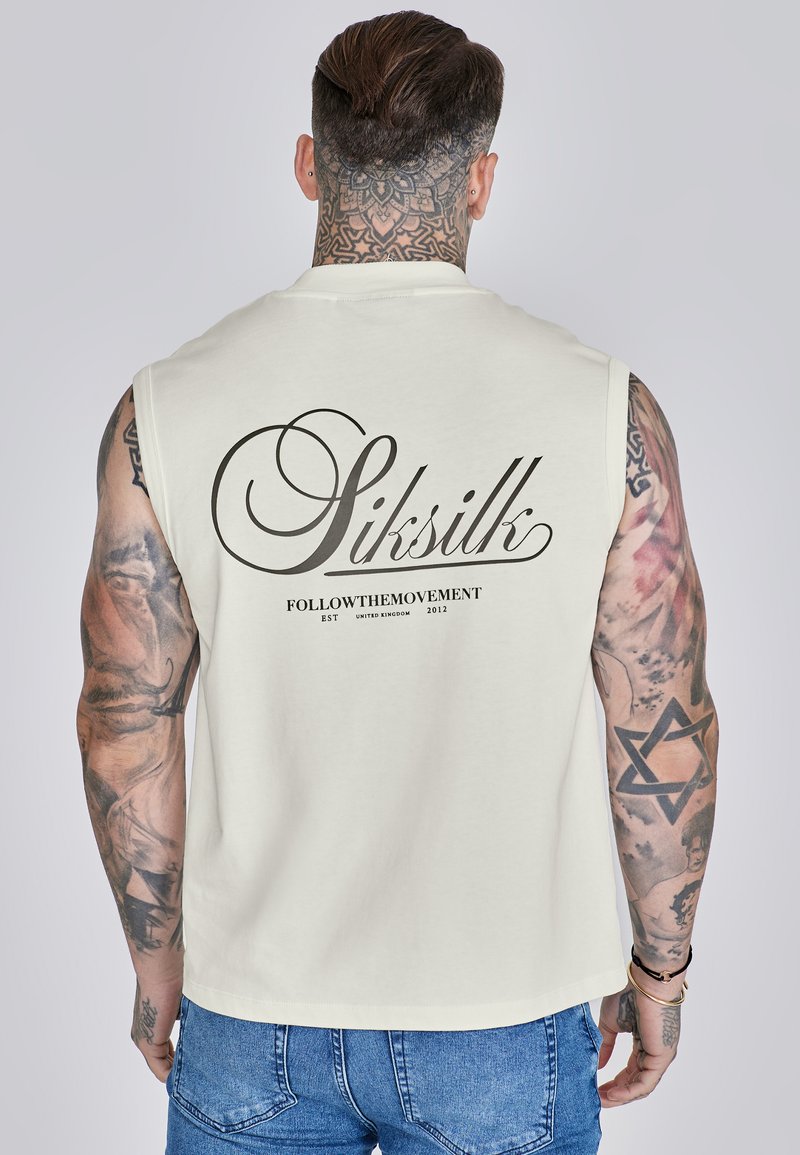 Chemise sans manches beige clair avec un grand logo noir en script "Siksilk" au dos, présentant des bords nets et une coupe ample.