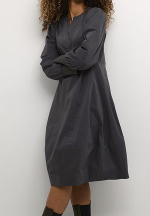 Frau mit lockigem Haar trägt ein lockeres, langärmeliges schwarzes Kleid und schwarze Stiefel und steht mit verschränkten Armen vor einer weißen Wand.