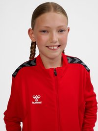 Fille souriante aux cheveux tressés portant une veste de sport rouge Hummel avec des bandes chevrons noires et grises sur les épaules.