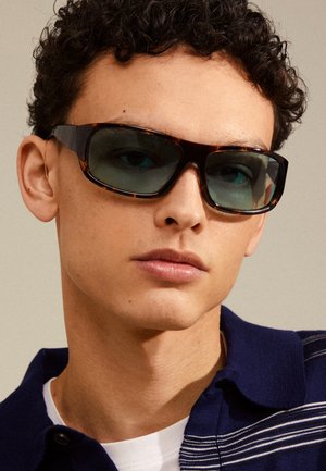 Occhiali da sole con montatura tartarugata, lenti scure e forma geometrica. Indossati da un modello con capelli ricci, su uno sfondo neutro.