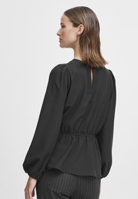 Blusa nera a maniche lunghe con vita aderente, dettaglio a occhiello sulla schiena e tessuto morbido. Presenta maniche arricciate per volume e movimento.