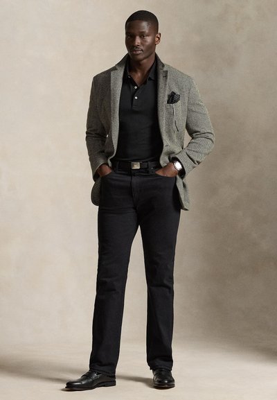 Mann trägt grauen Blazer, schwarzes Poloshirt, schwarze Hose, schwarze Lederschuhe und Armbanduhr, steht mit Händen in den Taschen vor einfachem Hintergrund.