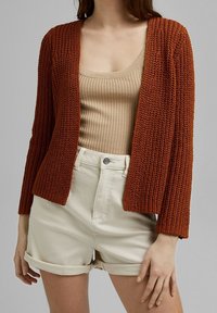 Rust-colored knitted cardigan with a relaxed fit and open front, paired with a beige ribbed tank top and light-colored rolled shorts.