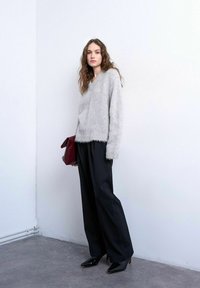 Femme aux cheveux ondulés portant un pull gris duveteux, un pantalon large noir, des bottes à talons noires, tenant une pochette rouge foncé contre un mur blanc.