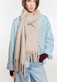 Beige ullsjal med fransade ändar, mjuk textur, svept runt en person, parat med en grön och vit randig skjorta.
