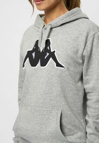 Grijze hoodie met een zwart grafisch logo op de voorkant. Het heeft een voorzak, een capuchon met koord en een zachte, gestructureerde stof.