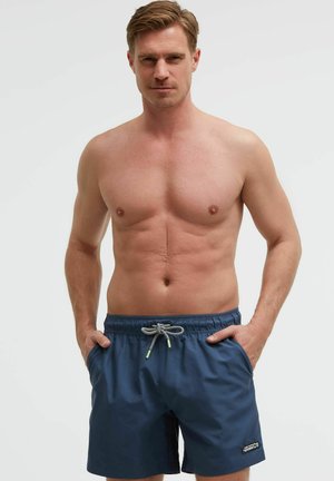 Hemdloser Mann mit kurzen blonden Haaren, der marineblaue Badeshorts trägt, mit den Händen in den Taschen stehend vor einem schlichten hellen Hintergrund.