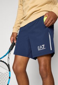Navy shorts made of lightweight fabric, featuring a grey accent stripe, elastic waistband, and EA7 logo on the front. Tennis ball held in pocket.