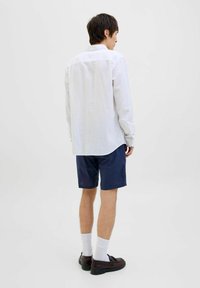Witte lange mouw blouse in de donkerblauwe shorts gestopt. De blouse heeft een button-down kraag; de shorts zijn knielang met een ontspannen pasvorm. Bruine schoenen en witte sokken.
