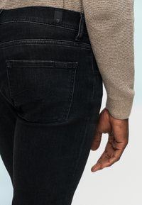 Κοντινό πλάνο ενός ατόμου που φορά μαύρο τζιν και μπεζ πλεκτό πουλόβερ, με έμφαση στην πίσω τσέπη και το δεξί χέρι που ακουμπά στο πλάι.