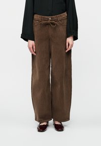 Isik, kellel on seljas pruunid laiad puuvillased püksid, millel on kangast vöös annab kauni lipsu, must pikkade varrukatega särk ja tumepunased lakknahast kingad.