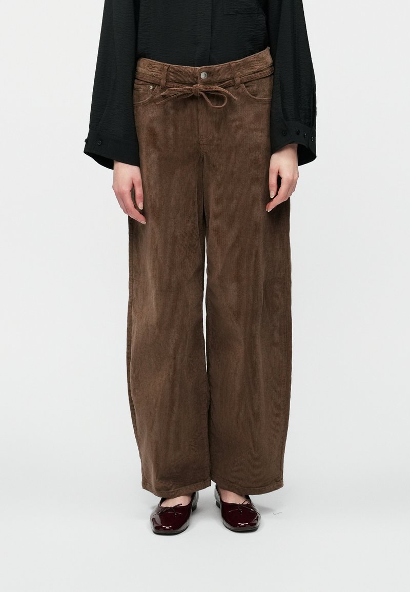 Isik, kellel on seljas pruunid laiad puuvillased püksid, millel on kangast vöös annab kauni lipsu, must pikkade varrukatega särk ja tumepunased lakknahast kingad.