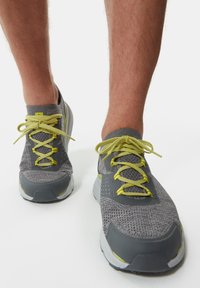 Zapatillas deportivas grises con parte superior de malla, cordones verde lima y suela de goma gris. Detalles destacables incluyen patrones texturizados en el costado.