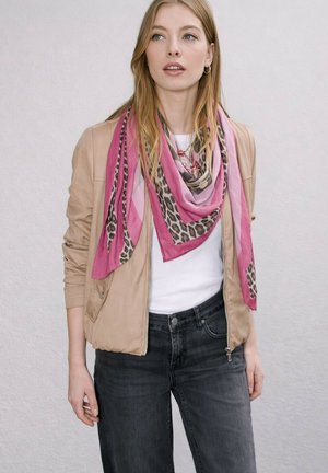 Vrouw die een beige jas, wit overhemd, zwarte spijkerbroek en een roze sjaal met luipaardprint draagt, staand tegen een lichte achtergrond.