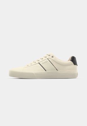 Cream-colored sneakers with a smooth texture, featuring black accents and branding. Classic lace-up design with a round toe and rubber sole.