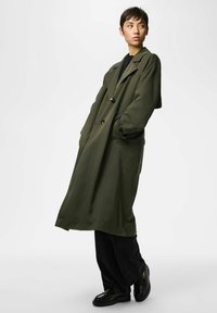 Noorik pikkade käistega oliivirohelise mantliga, mustade pükste ja läikivate mustade kingadega, seisab käed taskutes, vaatab küljele.