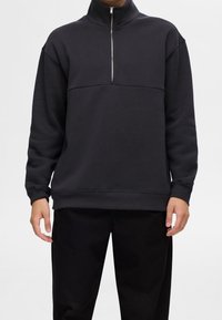 Svart kvartdragssäck med hög krage, med en slät textur och en horisontell söm över bröstet. Löst sittande design.