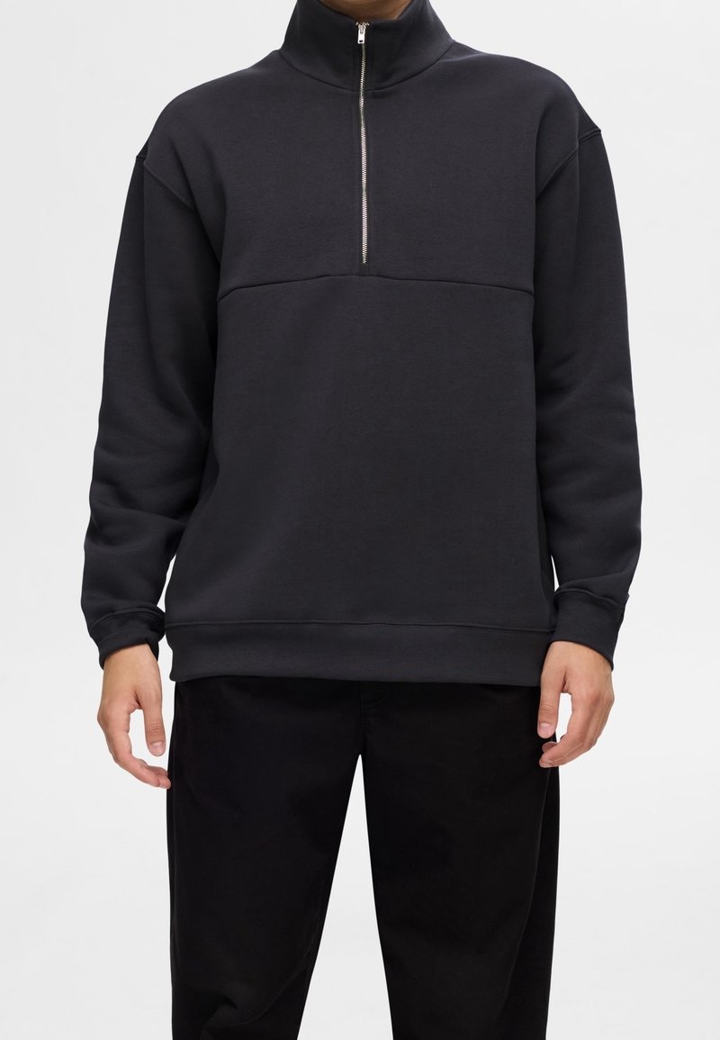 Svart kvartdragssäck med hög krage, med en slät textur och en horisontell söm över bröstet. Löst sittande design.