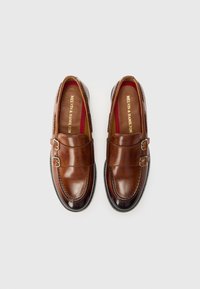 Brown leather loafers with double buckle detail, smooth texture, rounded toe, contrasting dark sole, and burgundy inner lining.
