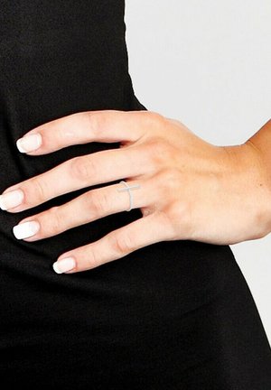 Main avec des ongles manucurés à la française, portant une fine bague en argent en forme de croix sur l'annulaire, sur un fond de tissu noir.