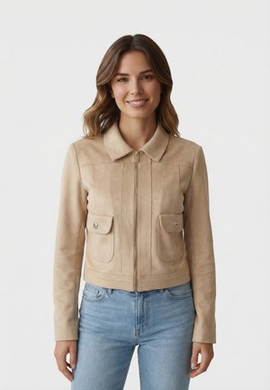 Femme souriante aux cheveux bruns ondulés portant une veste beige à fermeture éclair avec poches à boutons et un jean bleu clair, debout devant un fond uni.
