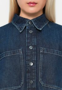 Donkerdenim overhemd met een kraag, sluiting met knopen aan de voorkant en twee borstzakken. Heeft contrasterende stiksels en zwarte knopen.