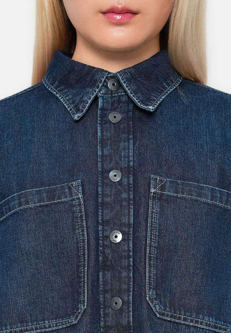 Mørk denimskjorte med krave, frontlukning med knapper og to brystlommer. Har kontrastsyninger og sorte knapper.