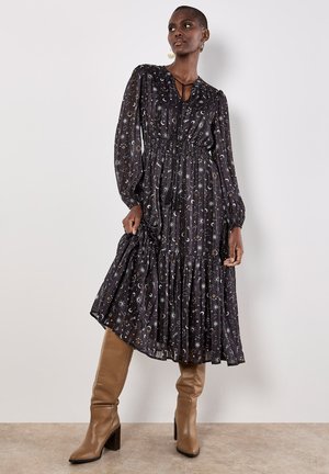 Zwarte maxi-jurk met hemelse patronen, geplooid design, gesnoerde taille en lange mouwen; gecombineerd met beige kniehoge laarzen.