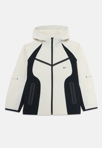 Veste à capuche beige clair avec des panneaux latéraux noirs, fermeture éclair à l'avant, poches zippées et petit logo swoosh noir sur la poitrine.