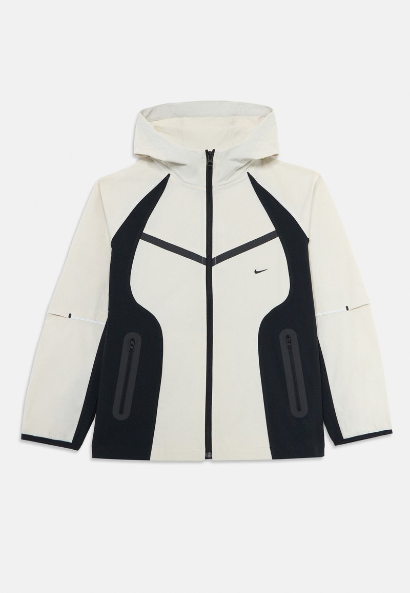 Veste à capuche beige clair avec des panneaux latéraux noirs, fermeture éclair à l'avant, poches zippées et petit logo swoosh noir sur la poitrine.