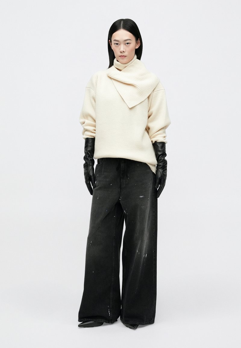 Crème gebreide trui met een sculpturale kraag, gecombineerd met loszittende zwarte denim broek met verfspetters, en zwarte lederen handschoenen.