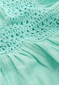 Turkise stof med detaljeret hækle-broderimønster og samlet, plisseret tekstur under lace-sektionen.