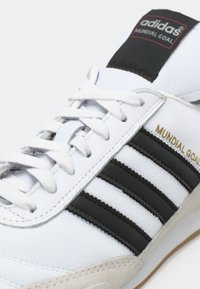 Białe buty piłkarskie Adidas Mundial Goal z czarnymi paskami, białymi sznurowadłami i beżowym detalem na podeszwie, z widocznym logo marki i nazwą modelu.