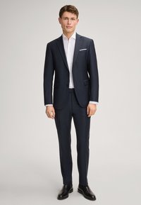 Marineblauwe kostuum met een op maat gemaakte pasvorm, enkele knoopsluiting, wit overhemd en zwarte leren schoenen. Bevat gestructureerde stof en zijzakken.