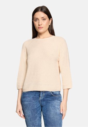 Woman with long brown hair wearing a light beige textured sweater with three-quarter sleeves and blue denim jeans, facing forward with a neutral expression.