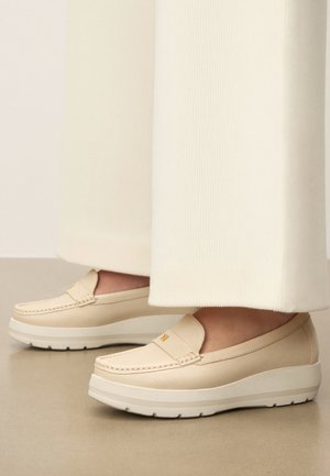 Bézsszínű bőr platform mokaszin, krémszínű bőszárú nadrággal viselve, sima padlón állva.