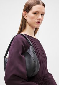 Čierna kožená kabelka s zakriveným dizajnom, s textúrovaným povrchom. Kabelku nesie na pleci osoba v fialovom topu.